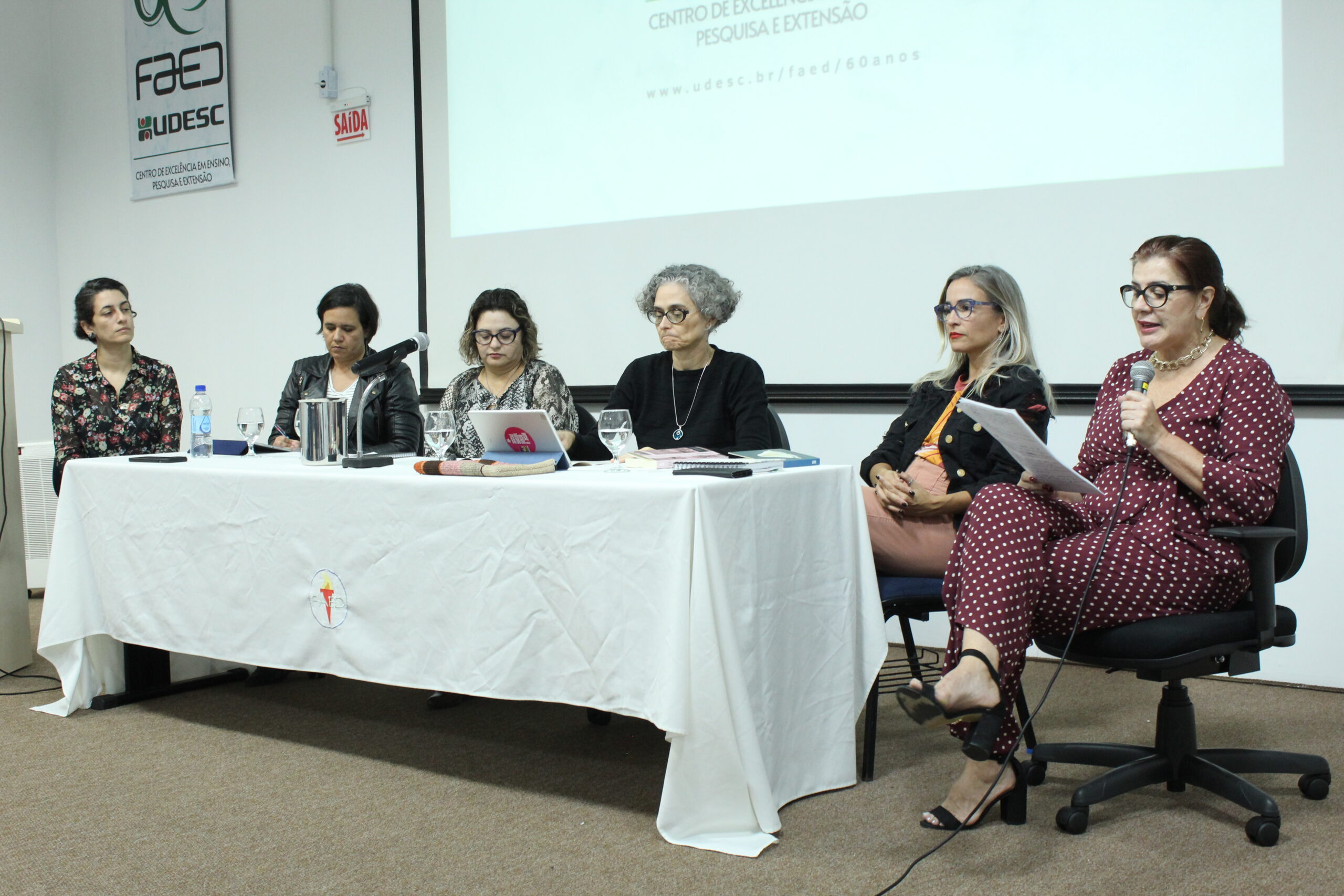 Seis mulheres sentadas atrás de uma mesa. Ao fundo, um slide show com apresentação.